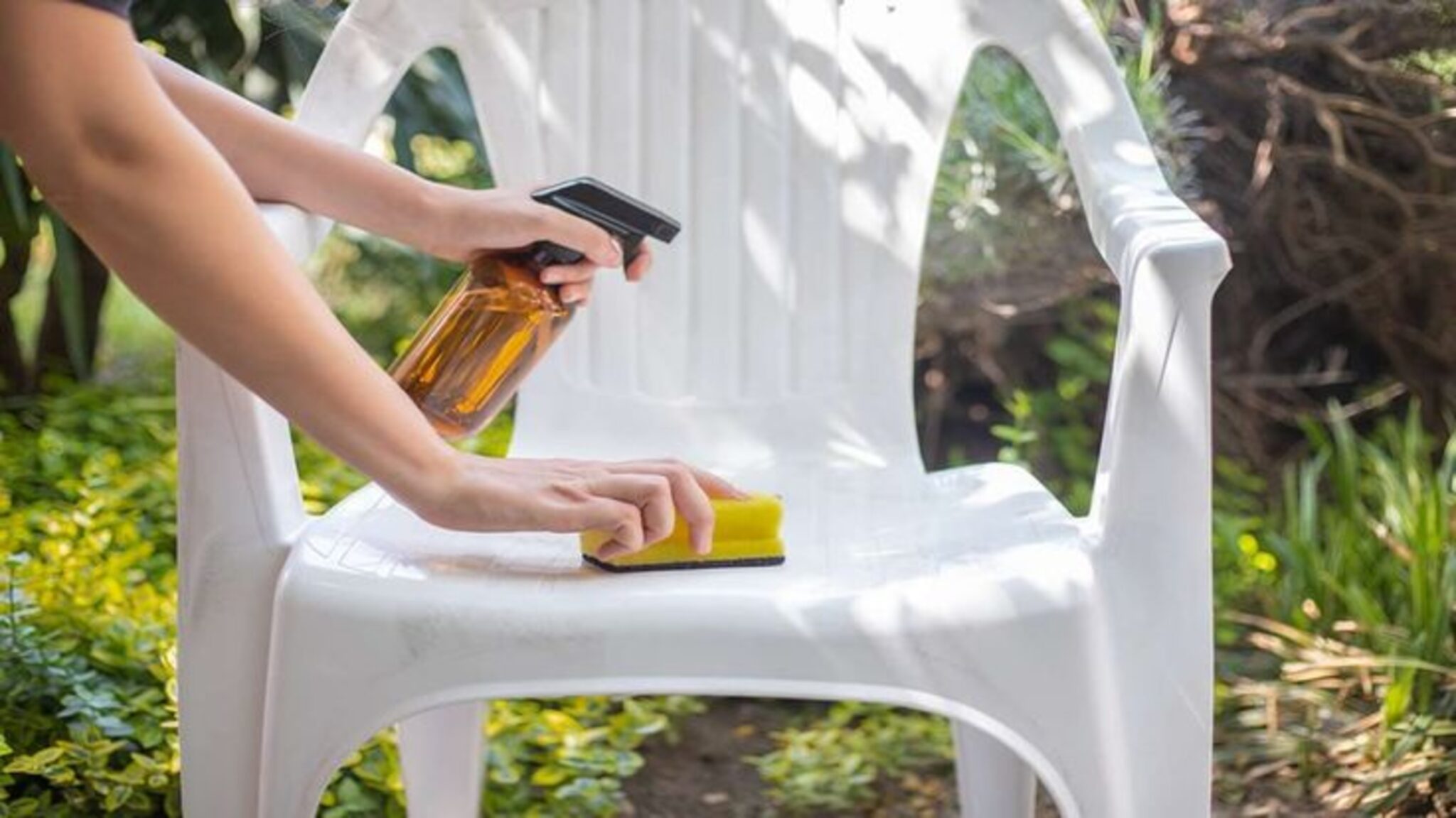 Person cleaning a white plastic chair with a spray bottle and sponge outdoors.