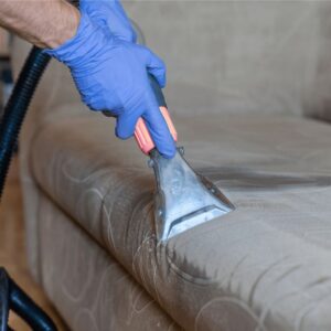 Person wearing gloves using an upholstery extractor to clean a fabric sofa seat.